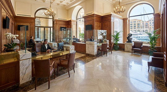 Elegant Monaco private bank interior with reception desks, chandeliers, and arched windows overlooking Port Hercule marina.
